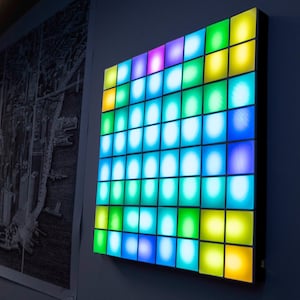 Könnte beinhalten: Ein quadratisches, an der Wand montiertes Lichtpaneel mit einem Raster aus beleuchteten Kacheln in leuchtenden Blau-, Grün-, Gelb- und Violetttönen. Das Display besteht aus mehreren einzelnen quadratischen Segmenten, die ein sanftes Licht abgeben.