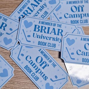 Op de afbeelding: Lichtblauwe rechthoekige tickets met afgeronde hoeken, verspreid op een houten oppervlak. De tickets bevatten de tekst "A Member of the Briar University Book Club" en "Off Campus Book Club". Elk ticket heeft een hockeystick en een hart.