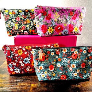 May include: Four floral print cosmetic bags in various colors: black, pink, red, and teal. Each bag features a different floral pattern with a matching tassel zipper pull. The bags are arranged on a wooden surface.