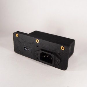 Pode incluir: Um módulo de alimentação retangular preto com uma entrada de energia IEC C14 embutida e um pequeno interruptor basculante com um símbolo I/O. A placa frontal inclui três inserções de latão roscado dourado nos cantos e é fixada com quatro parafusos pretos.