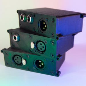 Puede incluir: Tres cajas electrónicas negras apiladas. Cada caja tiene múltiples puertos de entrada/salida, incluyendo conectores XLR y conectores jack de 6,35 mm. Las cajas están aseguradas con pequeños tornillos negros.