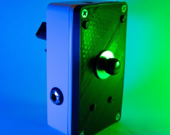 May include: A rectangular electronic device with a white and black casing. The device has a black knob, a small black port, and several screws. The background is a gradient of blue and green.