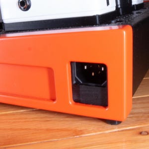 Könnte beinhalten: Nahaufnahme eines orangefarbenen und schwarzen elektronischen Geräts mit einem schwarzen Stromanschluss an der Unterseite.