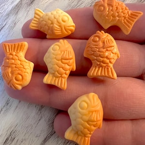 May include: Seven small, orange fish-shaped candies. The candies have detailed features, including fins, eyes, and scales. The candies are a light orange color and appear to be made of a hard candy or a similar material.