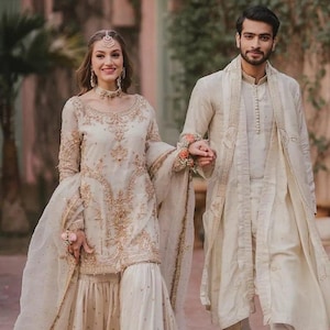 Puede incluir: Una pareja con atuendos tradicionales color crema, probablemente para una boda. La mujer lleva un vestido adornado con un tocado y joyas a juego. El hombre viste un conjunto coordinado. Ambos conjuntos presentan bordados dorados.