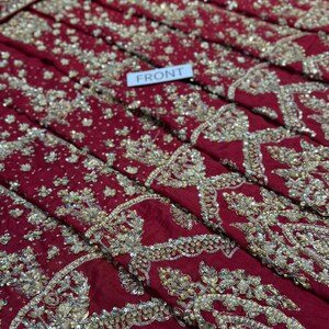 May include: Close-up of a deep red fabric with intricate gold embroidery and beadwork. The fabric is folded, revealing layers of detailed patterns. A small white tag reads "FRONT".