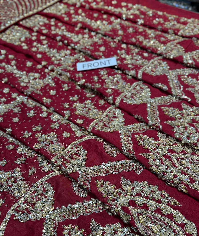 May include: Close-up of a deep red fabric with intricate gold embroidery and beadwork. The fabric is folded, revealing layers of detailed patterns. A small white tag reads "FRONT".