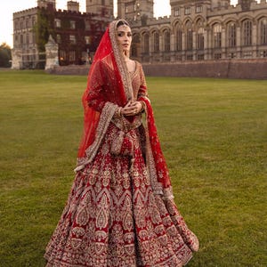 May include: A woman in a red and gold embroidered bridal gown stands in front of a castle. The gown features intricate patterns and a matching veil. The ensemble includes a small purse. The background is a green lawn and a stone castle.