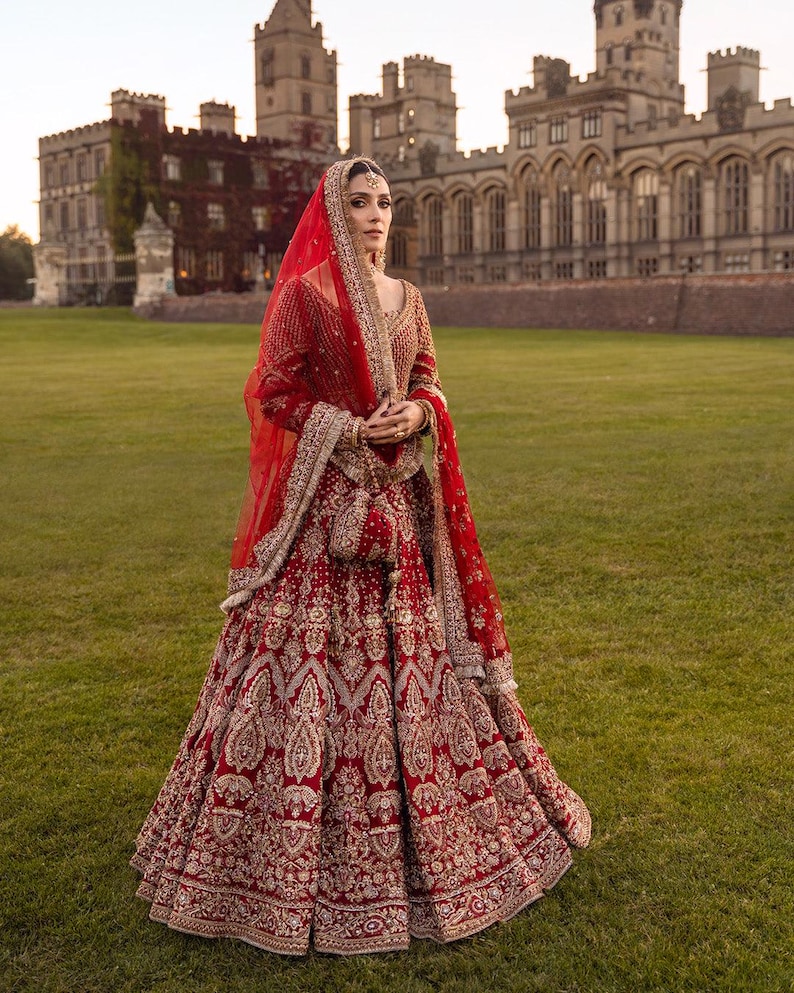 May include: A woman in a red and gold embroidered bridal gown stands in front of a castle. The gown features intricate patterns and a matching veil. The ensemble includes a small purse. The background is a green lawn and a stone castle.