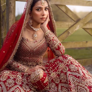 May include: A red and gold embroidered bridal gown with matching head covering and jewelry. The dress features intricate gold detailing and embellishments. The bride is wearing a necklace, earrings, and a headpiece.