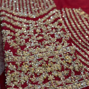 May include: Close-up of a deep red fabric with intricate gold embroidery. The embroidery features floral and geometric patterns, with a mix of sequins and beads. The design is dense and detailed, creating a luxurious texture.