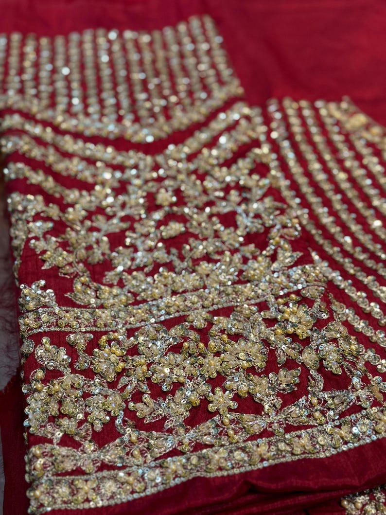 May include: Close-up of a deep red fabric with intricate gold embroidery. The embroidery features floral and geometric patterns, with a mix of sequins and beads. The design is dense and detailed, creating a luxurious texture.