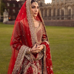 May include: A red bridal ensemble with gold embroidery and embellishments. The outfit includes a long-sleeved top, a skirt, and a sheer veil. The intricate details and rich colors suggest a traditional wedding garment.