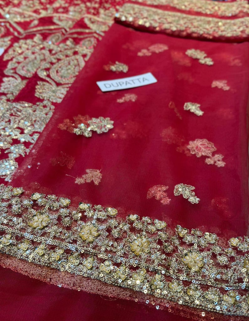 May include: A red dupatta with gold and silver embroidery. The dupatta has a delicate floral pattern and is embellished with sequins. The word "DUPATTA" is printed on a white tag. The fabric is sheer and lightweight.