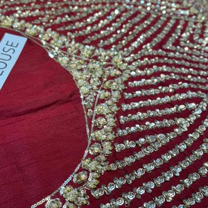 May include: Close-up of a burgundy fabric embellished with intricate gold and silver beadwork. The design features rows of sparkling beads and clusters of gold accents, creating a luxurious and ornate texture. The word "BLOUSE" is partially visible.