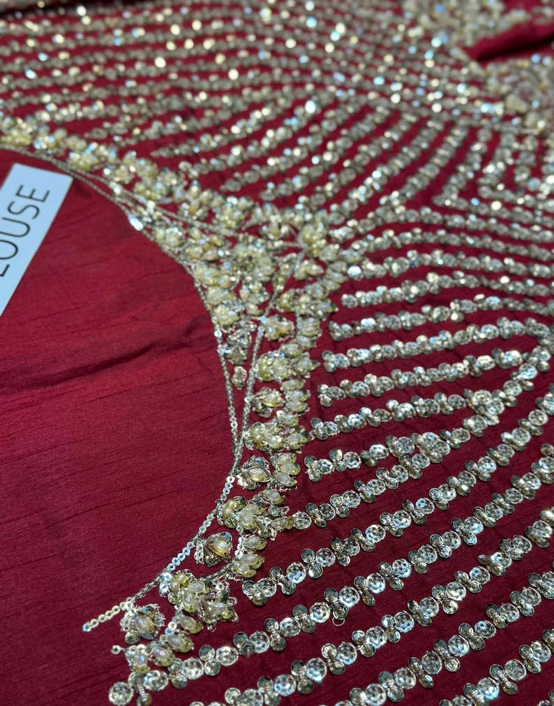 May include: Close-up of a burgundy fabric embellished with intricate gold and silver beadwork. The design features rows of sparkling beads and clusters of gold accents, creating a luxurious and ornate texture. The word "BLOUSE" is partially visible.