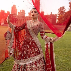 May include: A red and gold embellished bridal ensemble. The outfit includes a long-sleeved top and a full skirt, with intricate embroidery and beadwork. A sheer red veil is draped over the head and arms. The background features a castle and green lawn.