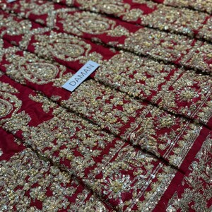 May include: Close-up of a section of red fabric heavily embroidered with gold-colored thread and sequins. The embroidery features floral and paisley patterns. A white tag with the word "DAMAN" is visible.