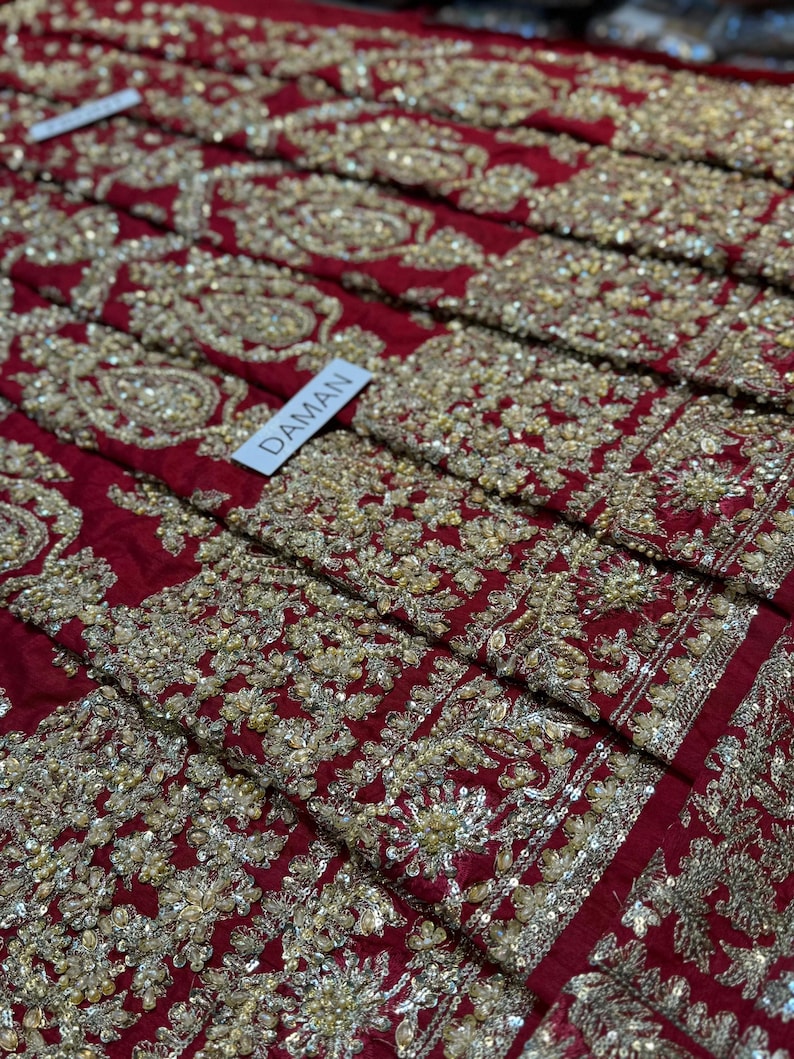 May include: Close-up of a section of red fabric heavily embroidered with gold-colored thread and sequins. The embroidery features floral and paisley patterns. A white tag with the word "DAMAN" is visible.
