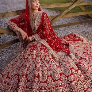 May include: A red bridal gown with intricate gold embroidery and embellishments. The gown features a full skirt and long sleeves, with a matching red head covering and shawl. The design includes detailed patterns and textures, creating a luxurious look.