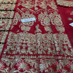 May include: Close-up of a red fabric with intricate gold embroidery and sequin embellishments. The design features floral and paisley patterns. A white label reads "BACK".