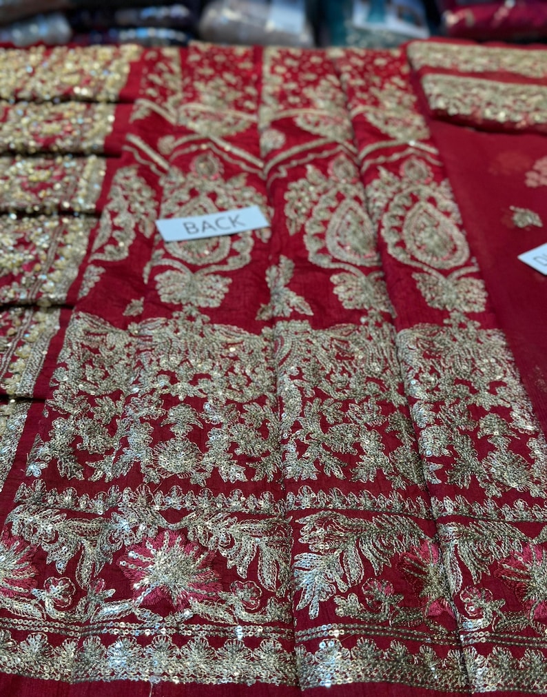 May include: Close-up of a red fabric with intricate gold embroidery and sequin embellishments. The design features floral and paisley patterns. A white label reads "BACK".