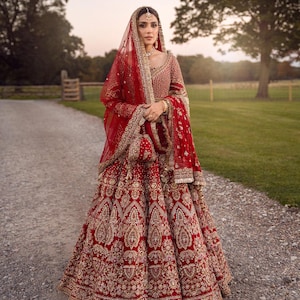 May include: A red bridal lehenga with intricate gold embroidery. The ensemble includes a matching dupatta, veil, and purse. The bride is wearing gold jewelry, including a headpiece and earrings. The dress is floor-length and features a detailed, ornate design.