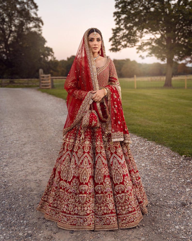 May include: A red bridal lehenga with intricate gold embroidery. The ensemble includes a matching dupatta, veil, and purse. The bride is wearing gold jewelry, including a headpiece and earrings. The dress is floor-length and features a detailed, ornate design.
