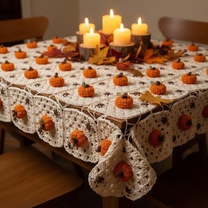 Puede incluir: Un mantel de ganchillo color crema con un patrón cuadrado repetitivo, cada uno adornado con una pequeña calabaza de lana naranja tridimensional. En el centro de la mesa hay cuatro velas de pilar encendidas rodeadas de hojas de otoño.