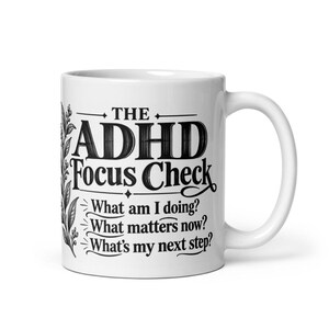 May include: A white ceramic mug featuring black text that reads THE ADHD Focus Check, followed by the questions: What am I doing? What matters now? What's my next step? A black botanical illustration of leaves runs vertically along the left side.