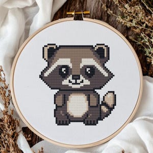 Puede incluir: Un patrón de punto de cruz de un lindo mapache con pelaje marrón, vientre blanco y nariz negra. El mapache está de pie sobre un fondo blanco.
