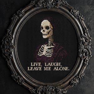 Op de afbeelding: Een zwart-witte kruissteekborduursel in een sierlijk zwart ovaal frame. De borduursel toont een skelet met een paars shirt en de tekst "Live, Laugh, Leave Me Alone."