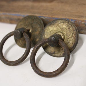 May include: Two circular brass drawer pulls featuring a weathered, dark patina. Each pull consists of a round, engraved backplate with a floral-inspired pattern and a hinged, dark-toned metal ring handle attached to the center of the plate.