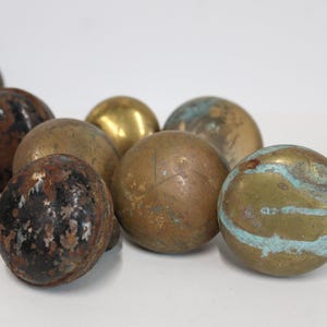 May include: A collection of antique brass spheres with varying patinas. Some spheres have a golden hue, while others display shades of brown, black, and turquoise. The spheres are arranged on a white surface, showcasing their aged textures.