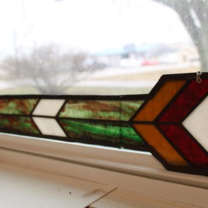 May include: Stained glass arrow with a black border. The arrow features a design with red, orange, white, green, and purple glass panels. It is hanging in front of a window.