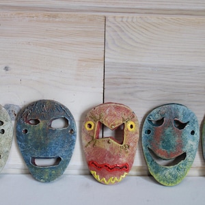 May include: Five handcrafted ceramic masks with expressive faces. Each mask features a unique color palette, including green, blue, and red, and distinct facial expressions. The masks are displayed against a light wood background.