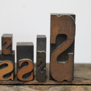 May include: Four rectangular wooden letterpress blocks of varying heights and widths. The blocks feature the letter S in different sizes and fonts, with a dark brown wood grain finish. The letters are carved in relief on the front faces.