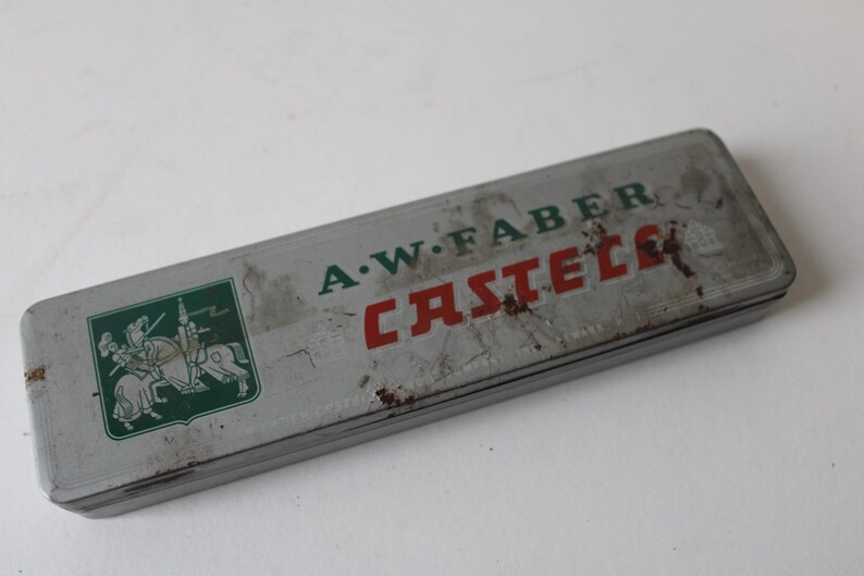 Pode incluir: Estojo de l&aacute;pis A.W. Faber Castell vintage. O estojo retangular de lata &eacute; cinza com letras verdes e vermelhas. Um emblema verde com cavaleiros a cavalo est&aacute; no lado esquerdo. O estojo mostra sinais de idade e desgaste.