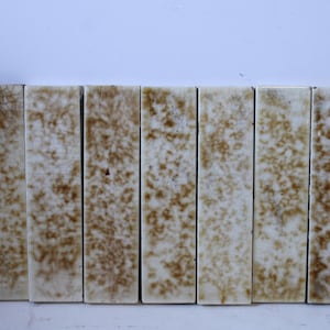 May include: A row of nine rectangular ceramic tiles featuring a glossy off-white base with a mottled, crackled brown glaze pattern. Each tile displays a unique, organic distribution of brown speckles and fine surface fissures across the surface.