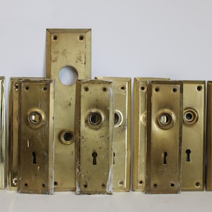 May include: A collection of antique brass door plates and hardware. The plates are rectangular with keyholes and circular knob holes. Some plates show signs of age and wear, with a warm, golden hue.