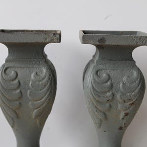 May include: Two gray, ornate architectural elements. Each has a square top, a bulbous midsection with a stylized leaf design, and a slender base. The pieces have a weathered appearance, suggesting they are vintage or antique.