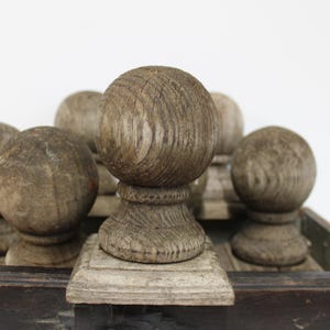 May include: A collection of six weathered wooden finials, each featuring a spherical top resting on a circular base and a square plinth. The wood displays a natural grain pattern with a muted, greyish-brown tone, arranged in a dark wooden tray.