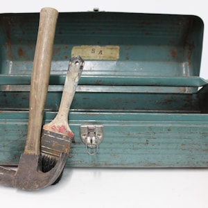 May include: An open, teal-colored metal toolbox with a hammer and paintbrush leaning against it. The toolbox has a latch and a label that reads "SA". The hammer has a wooden handle, and the paintbrush has a red and black bristle head.