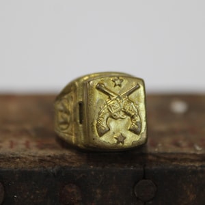 May include: A gold-colored ring with a rectangular face featuring crossed revolvers and stars. The ring has a detailed design on the sides, showcasing a vintage or antique style. The ring is set on a textured, dark surface.