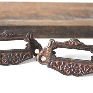May include: Two antique bronze drawer pulls with ornate floral and scrollwork designs. Each pull has a rectangular label holder and two mounting holes. The pulls have a distressed, aged appearance, suggesting they are vintage or vintage-inspired.