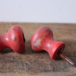 Puede incluir: Dos pomos de madera roja vintage con tornillos de metal oxidados. Los pomos tienen una forma redondeada con un acabado ligeramente desgastado, lo que sugiere edad y uso. Se encuentran sobre una superficie de madera r&uacute;stica.