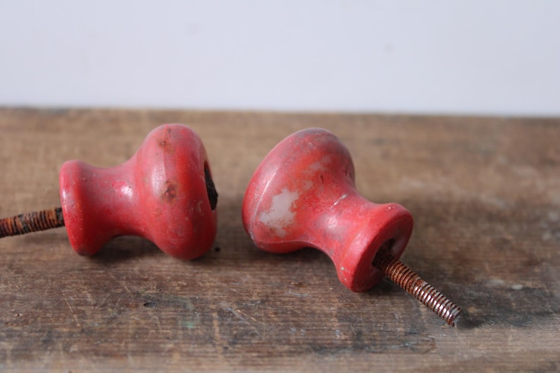 Puede incluir: Dos pomos de madera roja vintage con tornillos de metal oxidados. Los pomos tienen una forma redondeada con un acabado ligeramente desgastado, lo que sugiere edad y uso. Se encuentran sobre una superficie de madera r&uacute;stica.