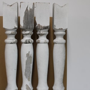May include: Four white, distressed wooden posts with decorative turned details. The posts have a weathered appearance with areas of exposed wood, suggesting a vintage or rustic aesthetic. Ideal for furniture or decor.
