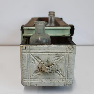 May include: An antique wooden box with a distressed white finish, featuring a carved floral design and a small handle. The box holds two vintage glass bottles. The box has a light green top and black trim.