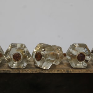 May include: A set of five clear, hexagonal glass door knobs arranged in a row. Each knob features a faceted, geometric design with a circular, rusted metal center component visible on the front face of the glass structure.